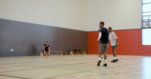Two players practicing pickup basketball on indoor hardwood court with bench and bags