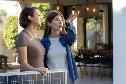 Young women holding solar panel, pointing and planning solar installation on sunny patio
