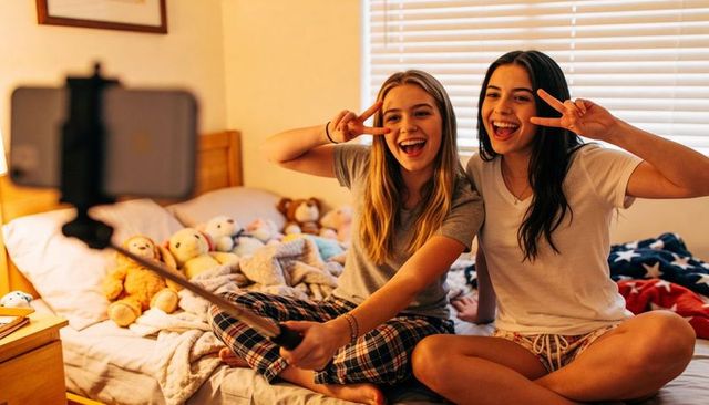 Teen girls taking joyful selfie on bed during cozy pajama sleepover with stuffed animals
