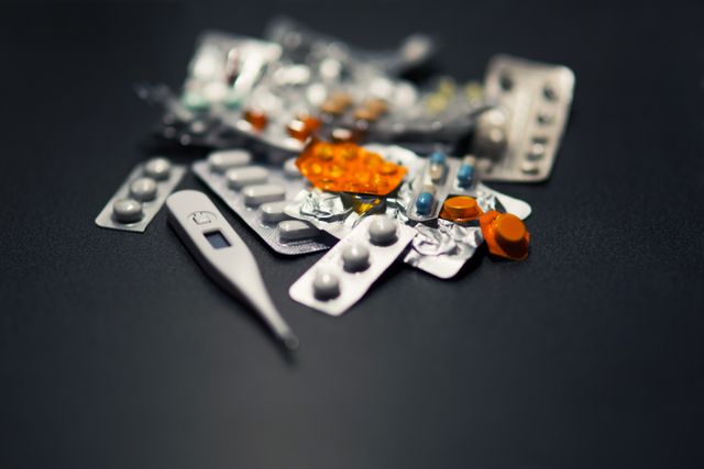 This image contains various blister packs of pills and tablets scattered next to a digital thermometer on a dark surface. It illustrates a theme of healthcare, medication, and treatment. Suitable for use in articles, brochures, or websites related to medical advice, pharmacies, illness management, or healthcare products.
