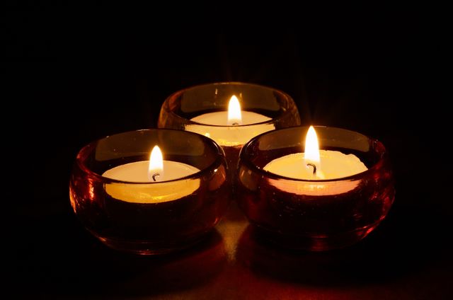 Three lit candles in round glass holders, casting a warm and tranquil light against a black background. Perfect for use in projects emphasizing relaxation, tranquility, meditation, romantic settings, or interior decor. Can also be ideal for themes focused on serenity, warmth, and ambiance creation.