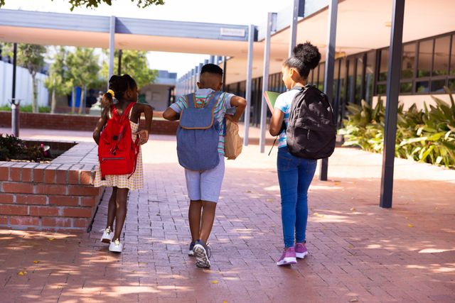 Three diverse children with backpacks walking towards an elementary school. Ideal for use in educational materials, back-to-school promotions, and articles on childhood and learning.