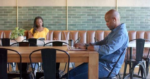 African man working on laptop in cafe with coffee cup and greenery, remote productivity