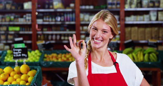 Cheerful grocery store clerk with a blonde braid wearing a red apron and white shirt in the produce section, gesturing an OK sign with her right hand. Fresh fruits and vegetables displayed in the background. Ideal for depicting friendly customer service, retail environments, and healthy food shopping experiences.