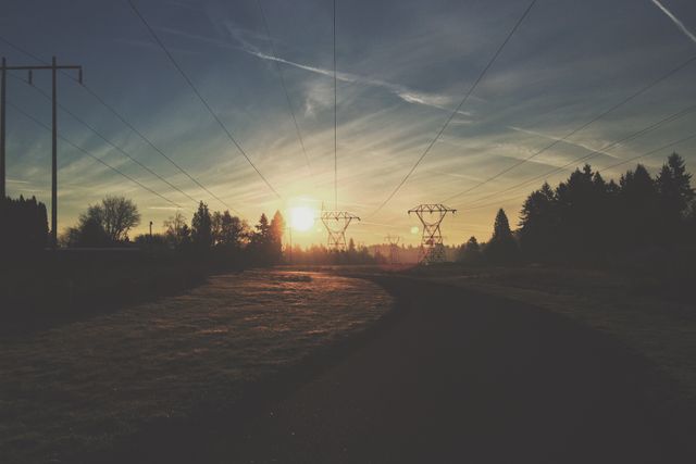 Beautiful scene showcasing a tranquil sunset over power lines and a country road in a rural area. Suitable for use in nature photography collections, energy industry presentations, and backgrounds for serene and peaceful themes.