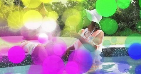 Chatting seniors wearing sunhats relaxing by backyard pool sipping drinks with pastel bokeh overlay