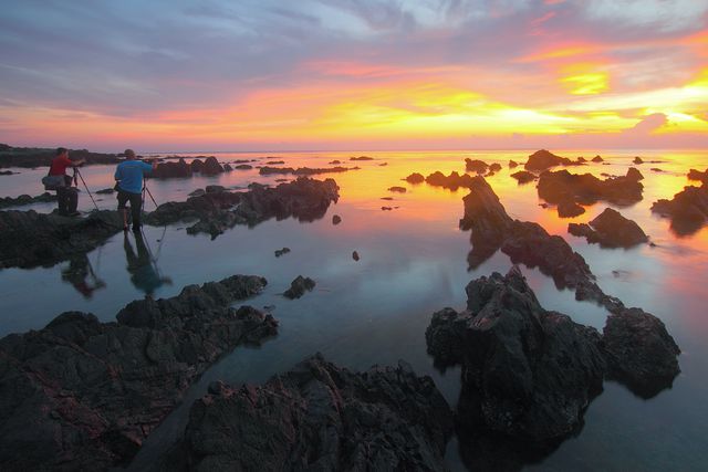 This image shows two photographers capturing a stunning sunset at a rocky beach during low tide. The dramatic, colorful sky reflects off the shallow water, creating a tranquil and serene scene. Perfect for use in travel brochures, nature blogs, photography tutorials, and relaxation content.