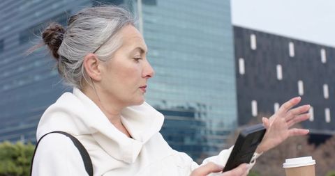 Senior woman checking smartphone and gesturing near takeaway coffee in urban plaza