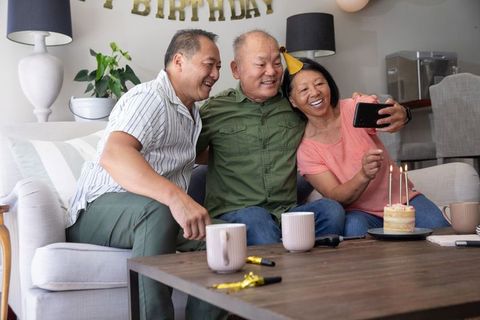 Asian family celebrating birthday at home taking selfie with cake, candles and party hat