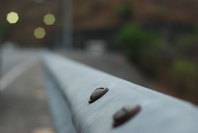 Metal guardrail up close on a roadside with blurred background. Suitable for use in materials on road safety, civil engineering, and infrastructure. Can also be used in nature vs. man-made environment topics, background textures, and photographic techniques highlighting depth of field and bokeh effects.