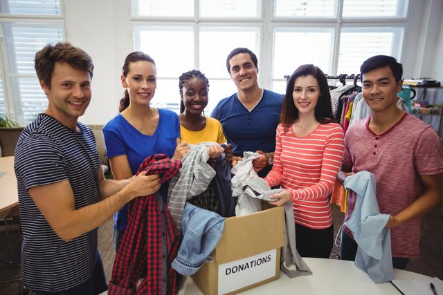 Diverse group of volunteers sorting clothes for donation in a bright room. They are smiling and holding various clothing items, standing around a box labeled 'Donations'. This image can be used for promoting volunteer work, charity events, community service projects, and nonprofit organizations. It highlights teamwork, diversity, and the spirit of giving back to the community.