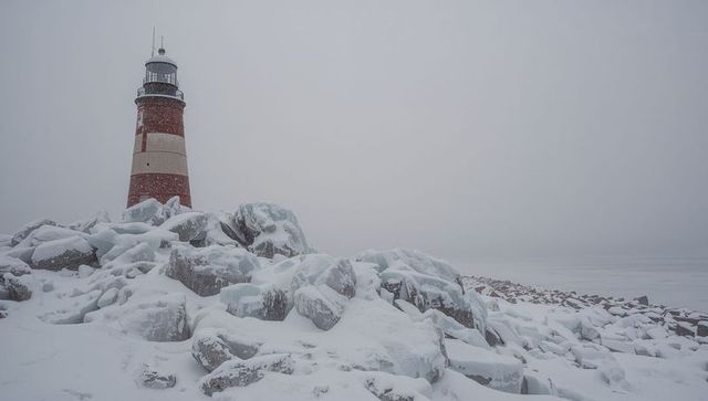 Red-and-cream lighthouse standing on snow-covered rocky coast in winter fog