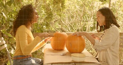 Friends carving pumpkins on sunlit porch table, scooping seeds and laughing in autumn
