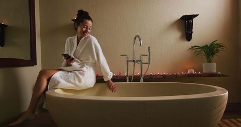 Woman in robe sitting on bathtub holding smartphone relaxing