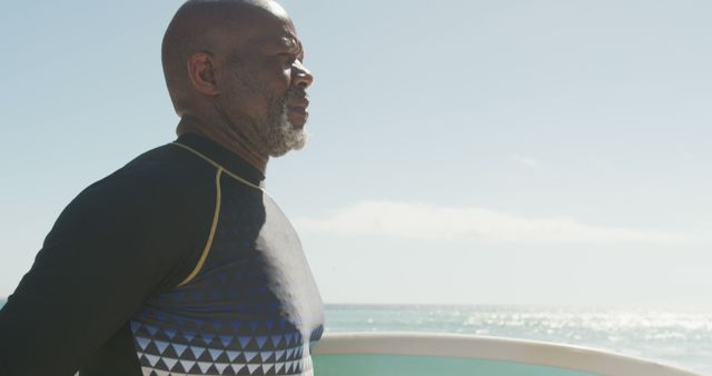 Senior African American man standing near ocean holding a surfboard, looking thoughtful. Perfect for themes of retirement, outdoor activities, lifestyle, and surf culture.