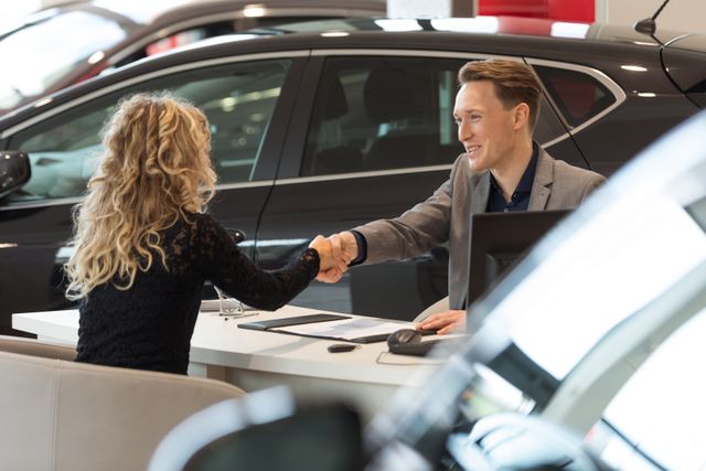 Smiling salesman doing handshake with female customer while sitting desk in car showroom