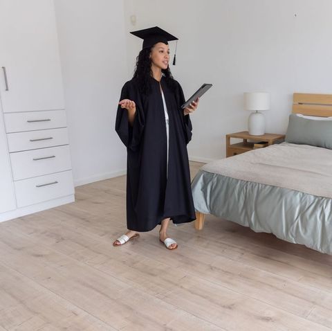 Teen female graduate wearing cap and gown giving virtual commencement speech in bedroom holding tabl