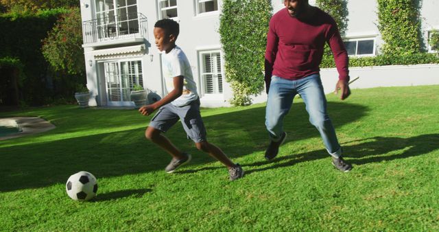 African american dad and son playing football together in the garden on a bright sunny day. fatherhood and love concept