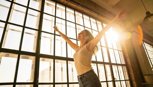 Young Woman Stretching into Sunlight in Industrial Loft with Paned Windows and Lens Flare