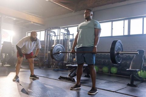 African american man lifting heavy barbell with trainer spotting in sunlit industrial gym