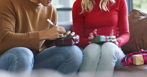 Couple wrapping gifts on couch writing personalized labels cozy holiday gift prep at home