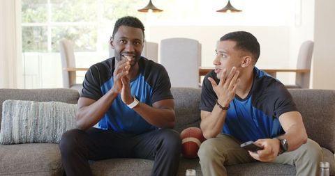 Diverse male friends cheering while watching football on couch with remote and ball