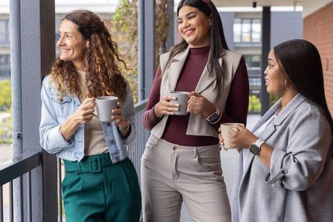 Diverse female colleagues chatting over coffee on office balcony during casual work break