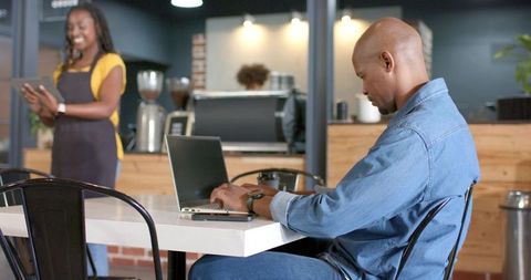 Denim-clad man typing on laptop in coffee shop while barista holding tablet, coworking