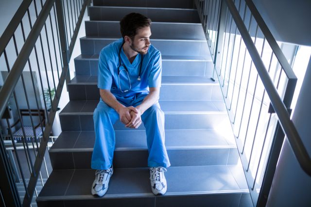 Male nurse in blue scrubs sitting on hospital staircase, appearing thoughtful and contemplative. Suitable for use in healthcare-related articles, mental health awareness campaigns, medical profession discussions, and stress management resources.
