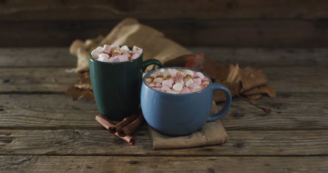 Two mugs of hot chocolate topped with marshmallows on rustic wooden table with autumn leaves and cinnamon sticks. Perfect for illustrating cozy, autumn, or seasonal themes. Ideal for use in blogs, holiday advertisements, and social media posts.