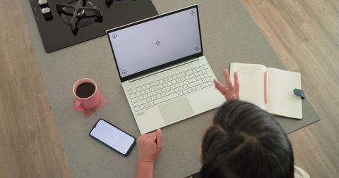 Mature woman working from home at kitchen island with laptop, coffee and notebook planning