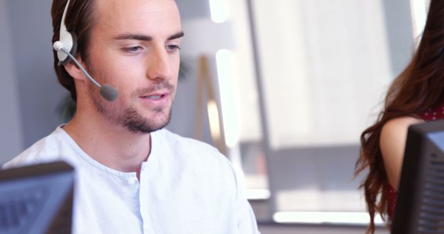 A young Caucasian man is working in a customer service environment, wearing a headset and focusing on a computer screen, with copy space. His professional demeanor suggests he is assisting a client or managing a support call.