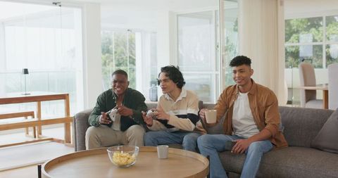 Multicultural young men playing video games on modern sofa, sharing chips and coffee mug