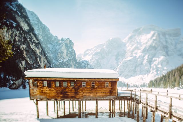 Depicts a rustic wooden cabin elevated on stilts against a backdrop of a frozen lake and snow-covered mountains. The serene winter setting suggests solitude and tranquil moments, making it ideal for promotions of winter travel, cabin rentals, outdoor adventure advertisements, and nature retreats. Perfect for use in travel blogs, magazines, postcards, or as a motivational poster showcasing serene natural beauty.