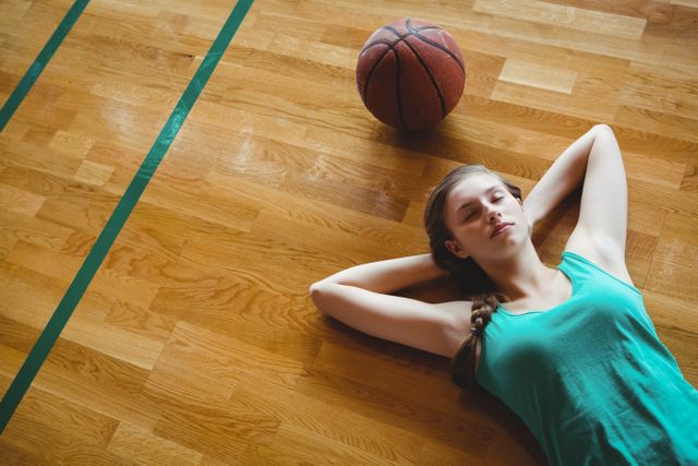 Young female basketball player taking a break and resting on the court. Ideal for use in sports-related articles, fitness blogs, and advertisements promoting athletic wear or sports equipment.