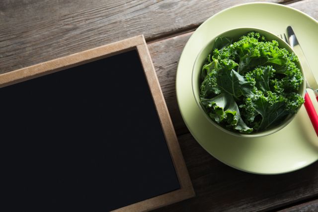 Fresh kale in a bowl placed on a plate next to a blank blackboard on a wooden table. Ideal for use in healthy eating promotions, organic food blogs, nutrition articles, and rustic kitchen decor themes.