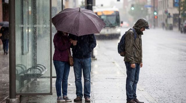 Couple sharing umbrella at rainy urban bus stop while lone hooded commuter standing apart
