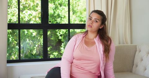 Pensive african american woman sitting on tufted sofa near window wearing pink activewear
