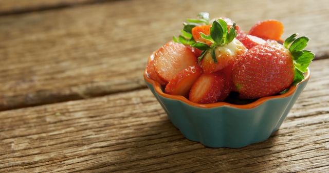 A blue ceramic bowl filled with fresh, ripe strawberries sits on a rustic wooden surface, with copy space. Vibrant in color, these strawberries offer a visual treat that suggests healthy eating and the freshness of summer produce.