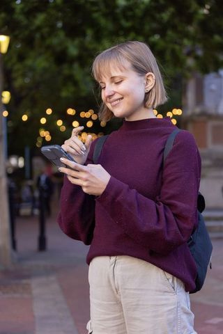 Young woman smiling and texting on smartphone at dusk with bokeh lights in urban plaza