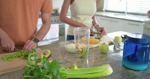 Diverse couple preparing green smoothie on granite countertop chopping avocado celery