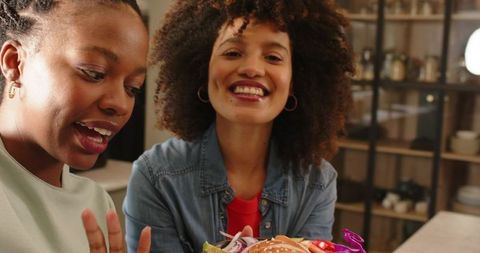 Smiling diverse women sharing colorful sesame burger in cozy modern kitchen scene