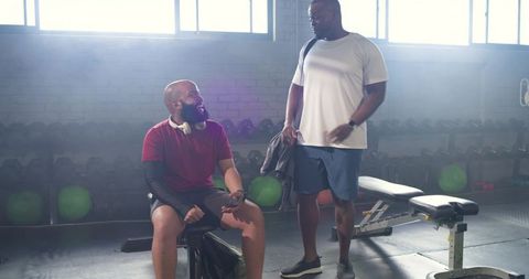African men resting and chatting after strength workout on bench with dumbbells and headphones