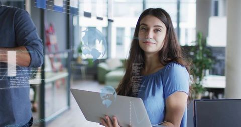 Woman holding laptop presenting digital globe overlays in modern open office for tech collaboration