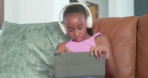 African american preteen girl wearing headphones using tablet on couch for learning