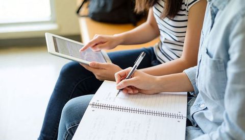 Students studying together using tablet while taking notes in spiral notebook on lap