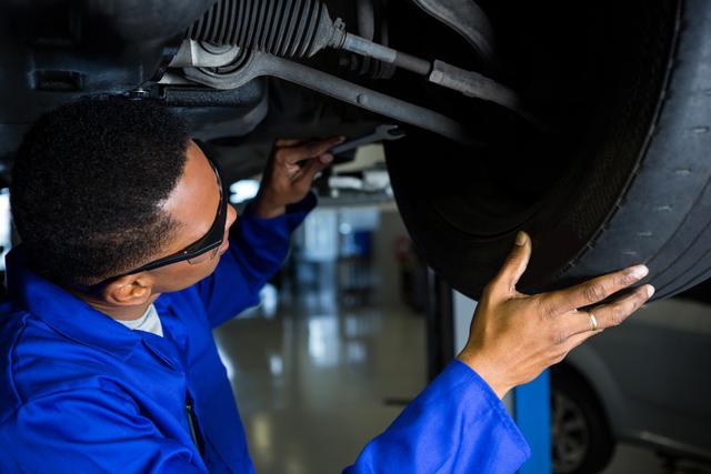 Mechanic in blue uniform inspecting car tire in an auto repair garage. Ideal for use in content related to automotive services, car maintenance, professional mechanics, and vehicle repair workshops.