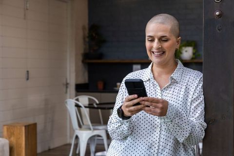 Smiling woman with shaved head checking smartphone while leaning on post in modern cafe