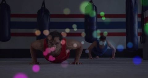 Man performing pushups in boxing gym with punching bags and colorful bokeh, duo