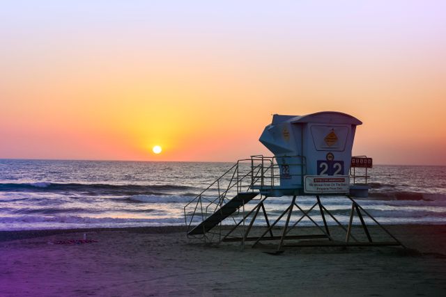 Lifeguard tower facing ocean during sunset with vibrant colors in sky. Waves gently rolling onto beach creating serene and tranquil atmosphere. Ideal for travel websites, summer destinations, seaside vacation promotions, and beach safety promotions.